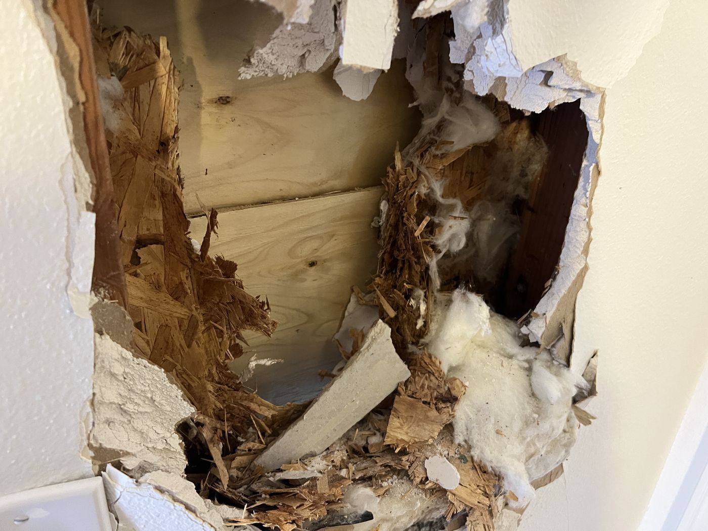 A damaged interior wall with a large hole exposing wooden structural elements, insulation material, and broken drywall. The wood appears splintered and torn, suggesting forceful impact or water damage.
