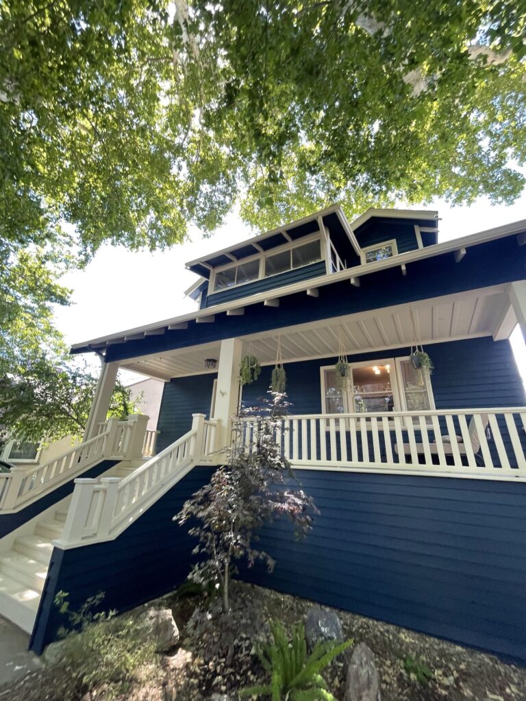 The front side of a blue two-story home with white trims. A white railing wraps around the front porch. a few small green plants are planted in front of the front porch.