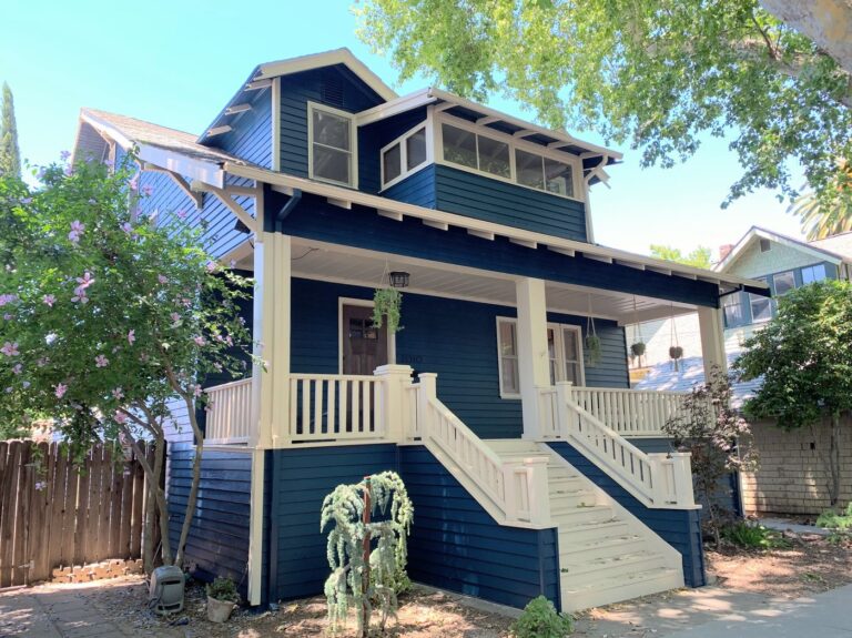 The front side of a blue two-story home with white trims. A white railing wraps around the front porch. a few small green plants are planted in front of the front porch.