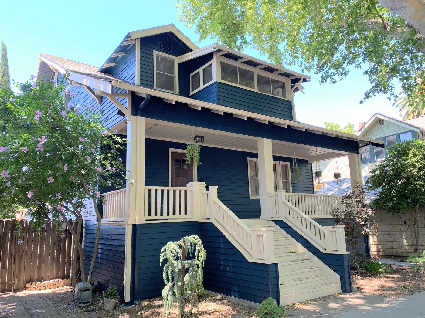 The front side of a blue two-story home with white trims. A white railing wraps around the front porch. a few small green plants are planted in front of the front porch.