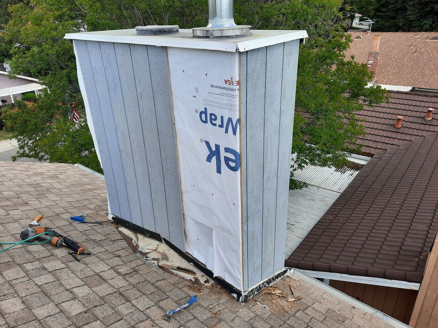 A rooftop chimney with white and gray vertical panel siding, surrounded by brown roof shingles. The base of the chimney shows exposed flashing with some sealant, ongoing maintenance work. A residential neighborhood and trees are visible.