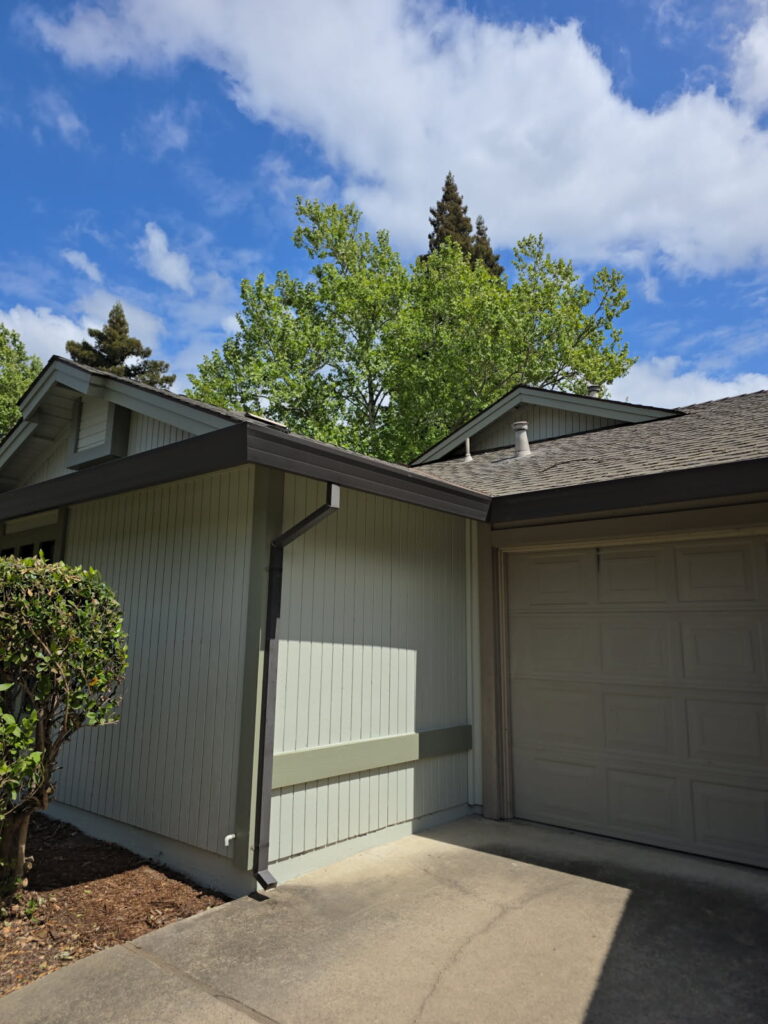 home exterior with light green vertical siding and dark trim, featuring a garage door and an angular downspout connected to the gutter system. The house is surrounded by green trees under a partly cloudy blue sky.